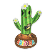 A plush toy resembling a saguaro cactus with a smiling face, adorned with white flowers and a 'Arizona' text base.