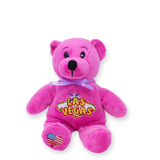 A pink plush bear with 'LAS VEGAS' embroidered on its chest, adorned with a purple bow and an American flag patch on its left paw.