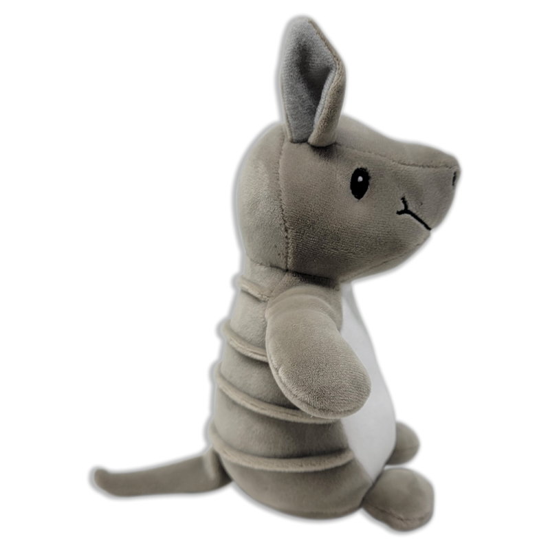 Side view of a gray plush armadillo with upright ears, stitched black eye, segmented body bands, white belly, and long tail on a transparent background.