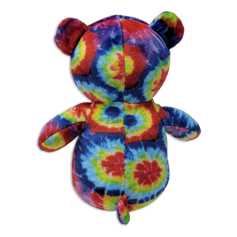 Back view of a tie-dye plush teddy bear with multicolored rainbow fabric, rounded ears, short arms, small tail, and visible center seam on a transparent background.
