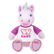 Front view of a pink plush unicorn with a rainbow horn and blue mane accents, wearing a white shirt with purple sleeves that reads “CONGRATS TO YOU! DAISY,” and sitting upright with pink padded feet.